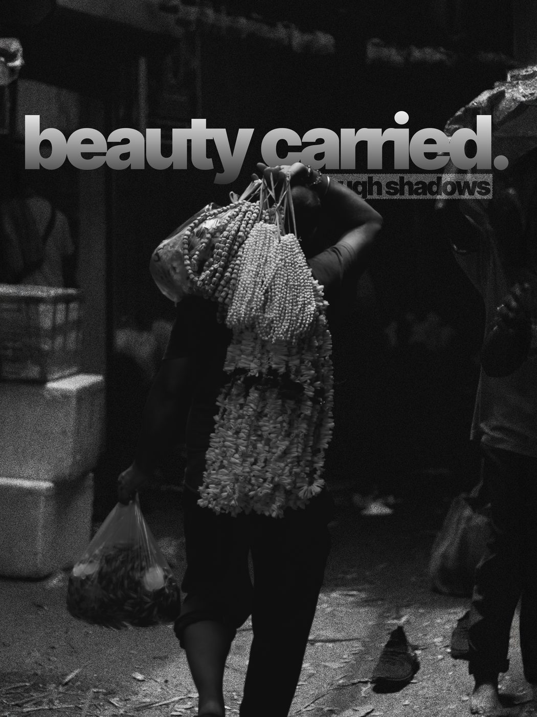 A woman carrying strung garlands on her head through a market, in black and white, captioned beauty carried through shadows